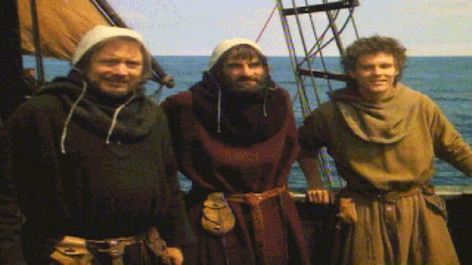 Três pessoas vestidas com roupas de estilo medieval — túnicas, cintos e capuzes — estão em pé no convés de um navio de madeira. Atrás delas aparecem cordas, mastros e parte da vela, com o mar ao fundo.