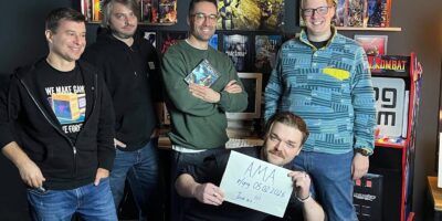 Cinco membros da equipe do GOG posam em uma sala decorada com memorabilia de videogames. Atrás delas há prateleiras cheias de caixas de jogos clássicos, como Gothic, Silent Hill 4: The Room e Breath of Fire. À direita, há uma máquina de arcade de Mortal Kombat. Uma pessoa em pé segura uma caixa de jogo; outra, sentada à frente, segura um cartaz escrito: ‘AMA — r/gog 05.02.2026 — Join us!!!’.
