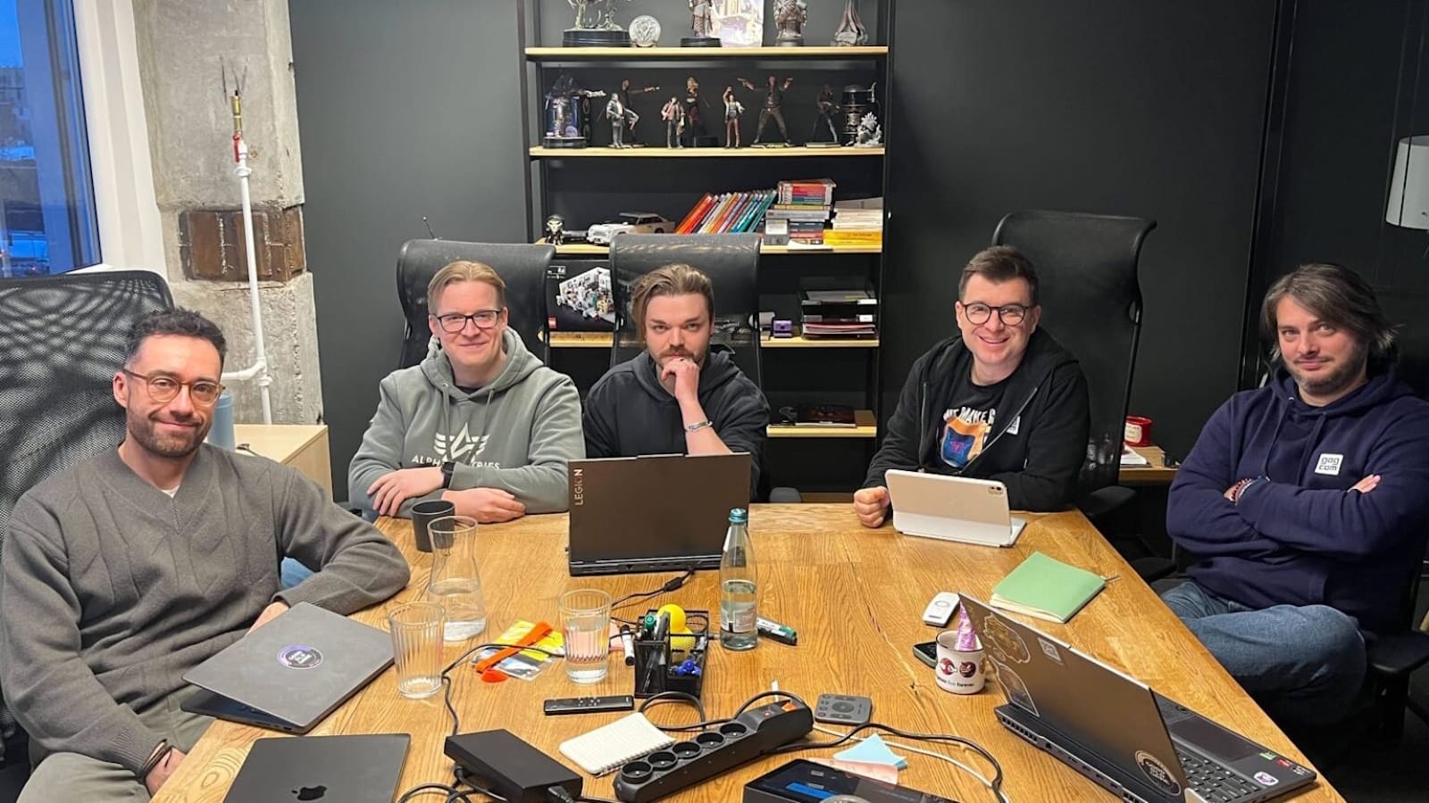 Cinco membros da equipe do GOG estão reunidas ao redor de uma mesa de madeira em uma sala de reunião moderna. Cada uma tem um laptop aberto, e a mesa está cheia de itens como cadernos, canetas, garrafa de água, copos, uma régua de energia e pequenos dispositivos eletrônicos. Atrás delas há uma estante preta com livros, figuras colecionáveis e objetos decorativos. 